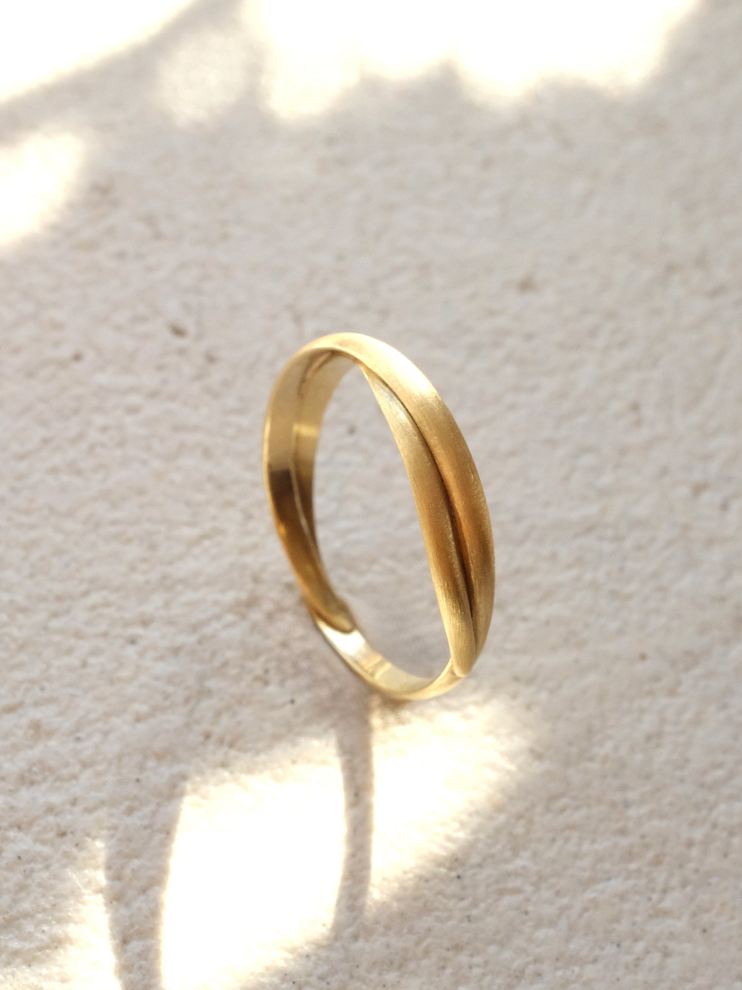 The same brushed gold ring band as the previous image shown upright from a three quarter view and in soft dappled light. Capturing the ring's layered form and the tapered ends tucking around and into the single wire strand at the ring's base.