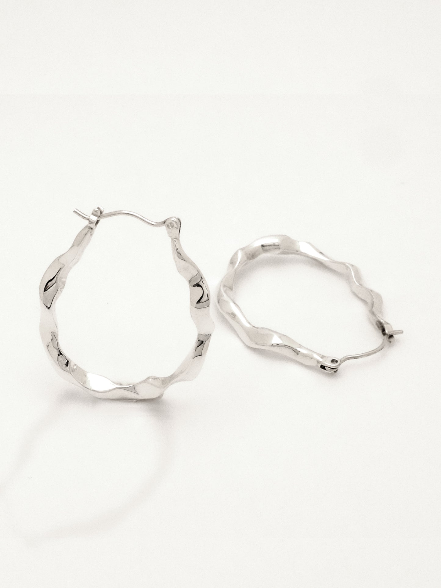 Close up of 2 sterling silver hoop earrings positioned to show the form from the side and three quarter view, conveying the undulating water like form as well as the hinged ear wire and catch.