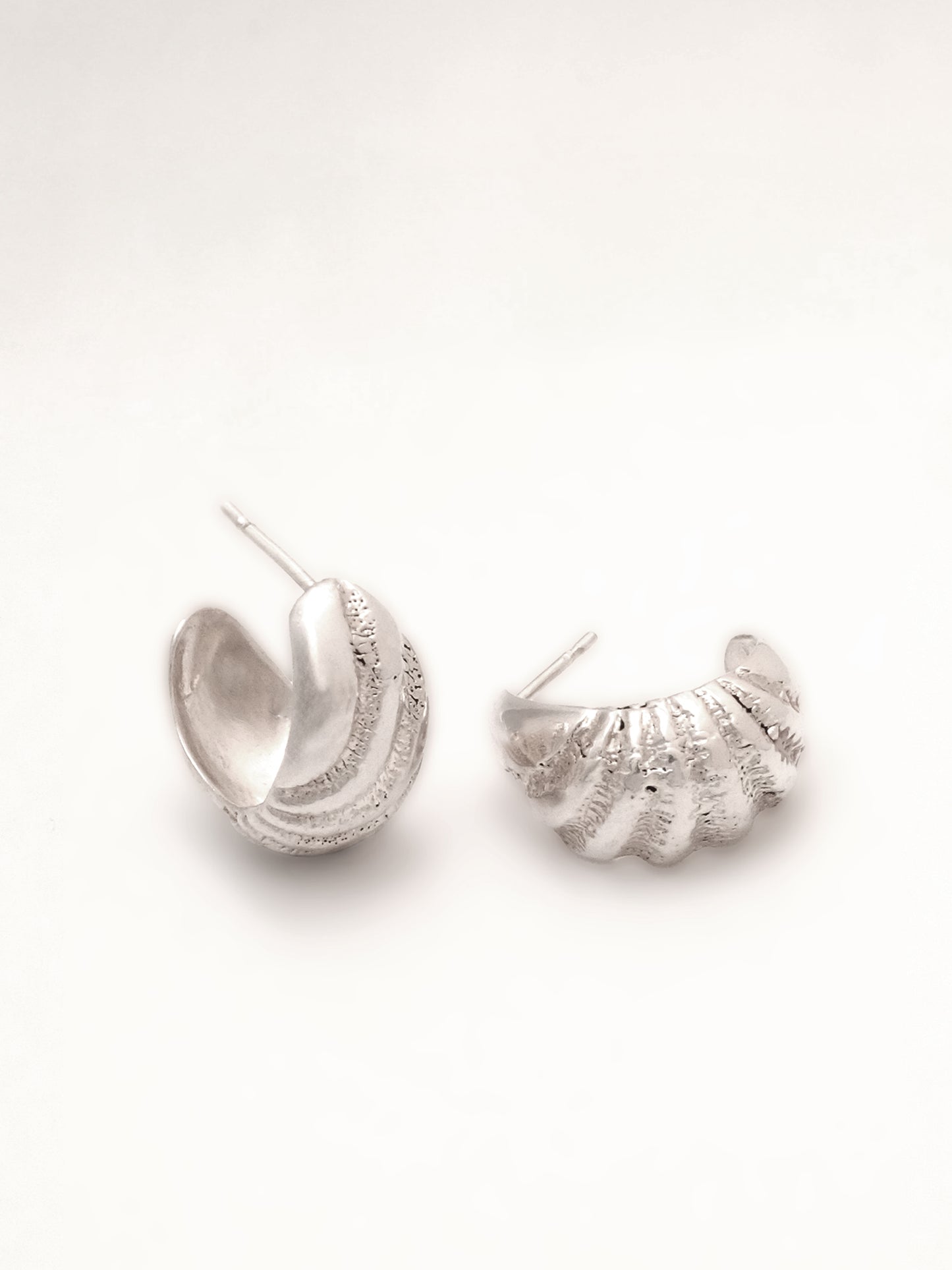 Close up of a set of sterling silver hoop earrings with posts. The pair are positioned to show the textural ridges the artist carved to mimic the texture of a sea-worn shell, the ridges radiating along the hoop's curved form.