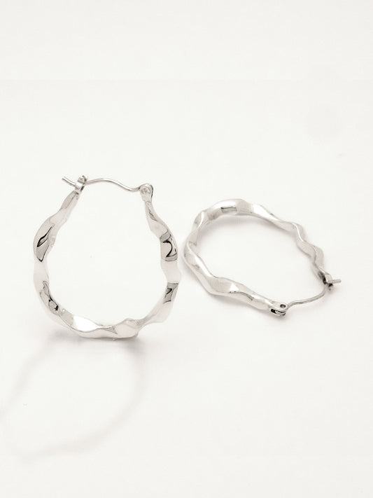 Close up of 2 sterling silver hoop earrings positioned to show the form from the side and three quarter view, conveying the undulating water like form as well as the hinged ear wire and catch. 