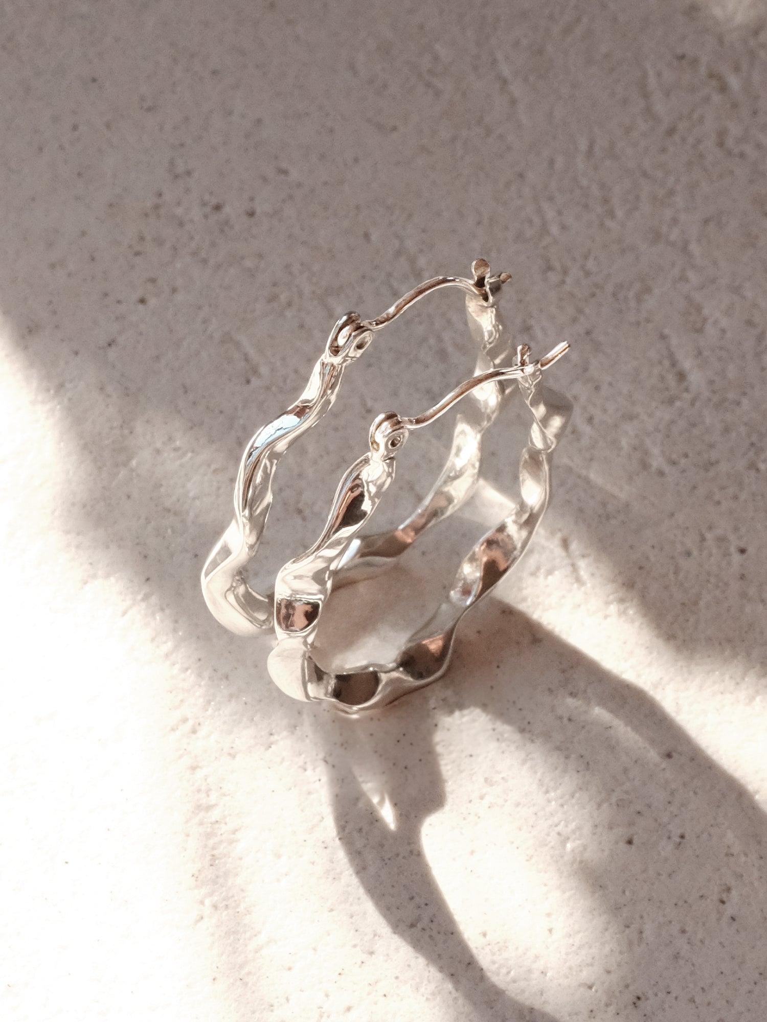 2 sterling silver hoop earrings positioned upright as a pair from a top down, three quarter view to give the of idea of their form as water trickling down. The dramatic light highlights how reflective the mirror surface is. 