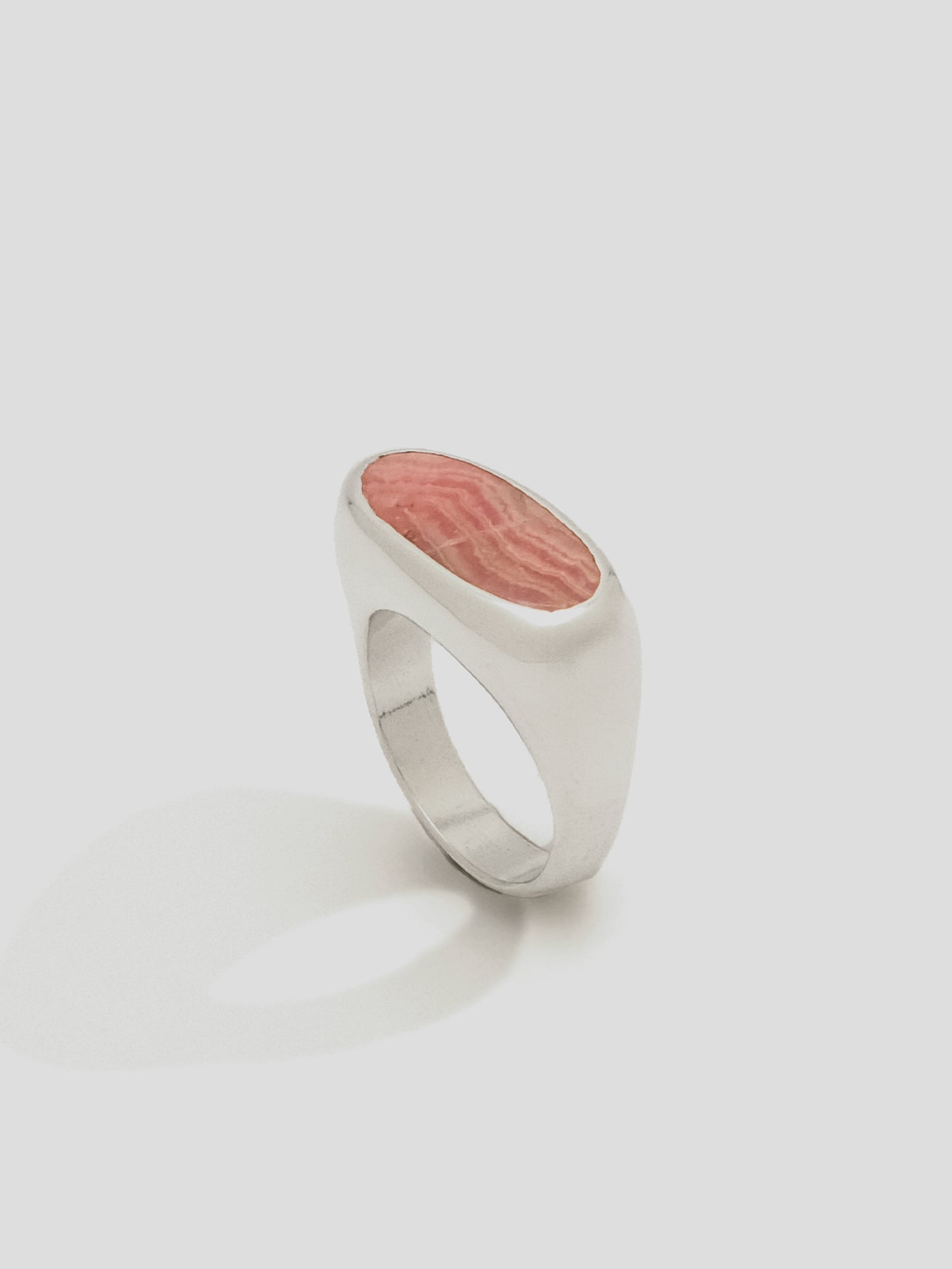 Close up of a Sterling Silver signet ring, with a pill shaped pink and rose toned stone with variegated striation throughout.   The ring is positioned upright to show it's softly curved profile tapering to the more narrow ring shank