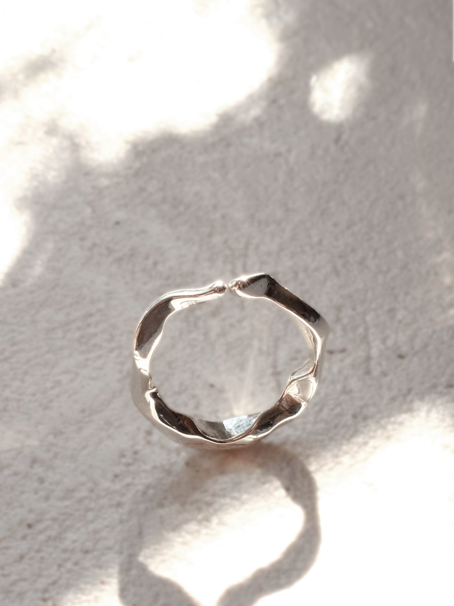 The same sterling silver ring as in the previous image, now positioned upright and captured in a beam of sunlight, which accentuates its undulating, water-like texture and mirror shine