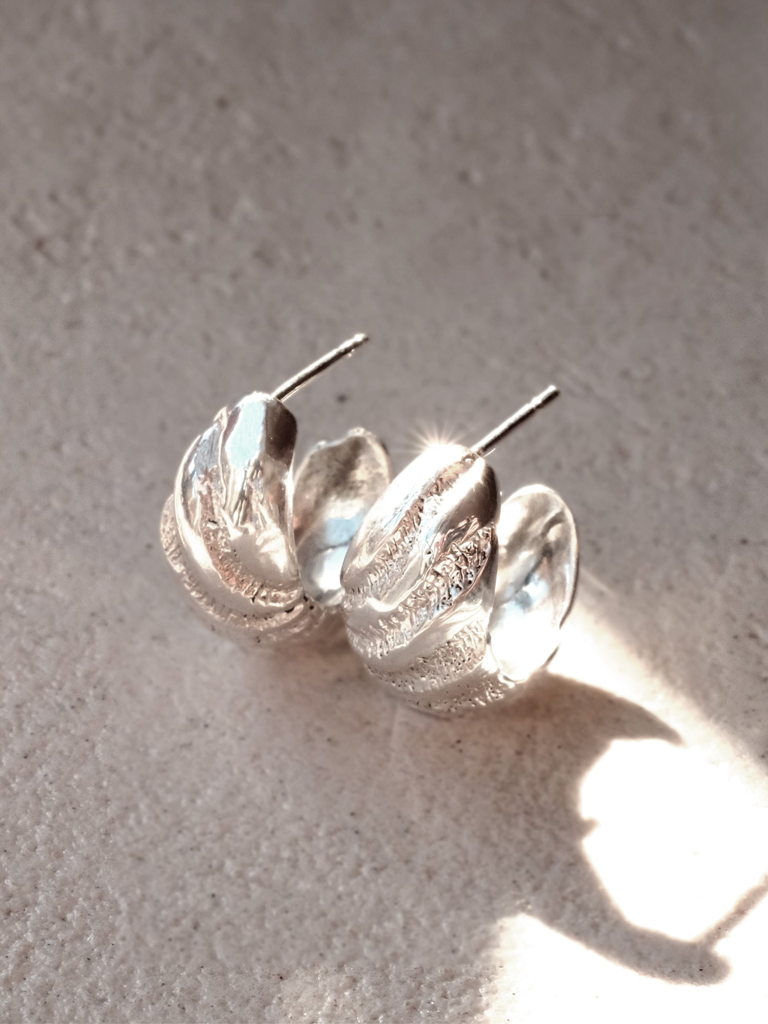 The same pair of  Sterling Silver hoop post earrings positioned upright and side by side, from a three quarter top down perspective, in a beam of sunlight to show the mirror finish, intricate texture and smooth clean finish of the interior of the hoop's form.