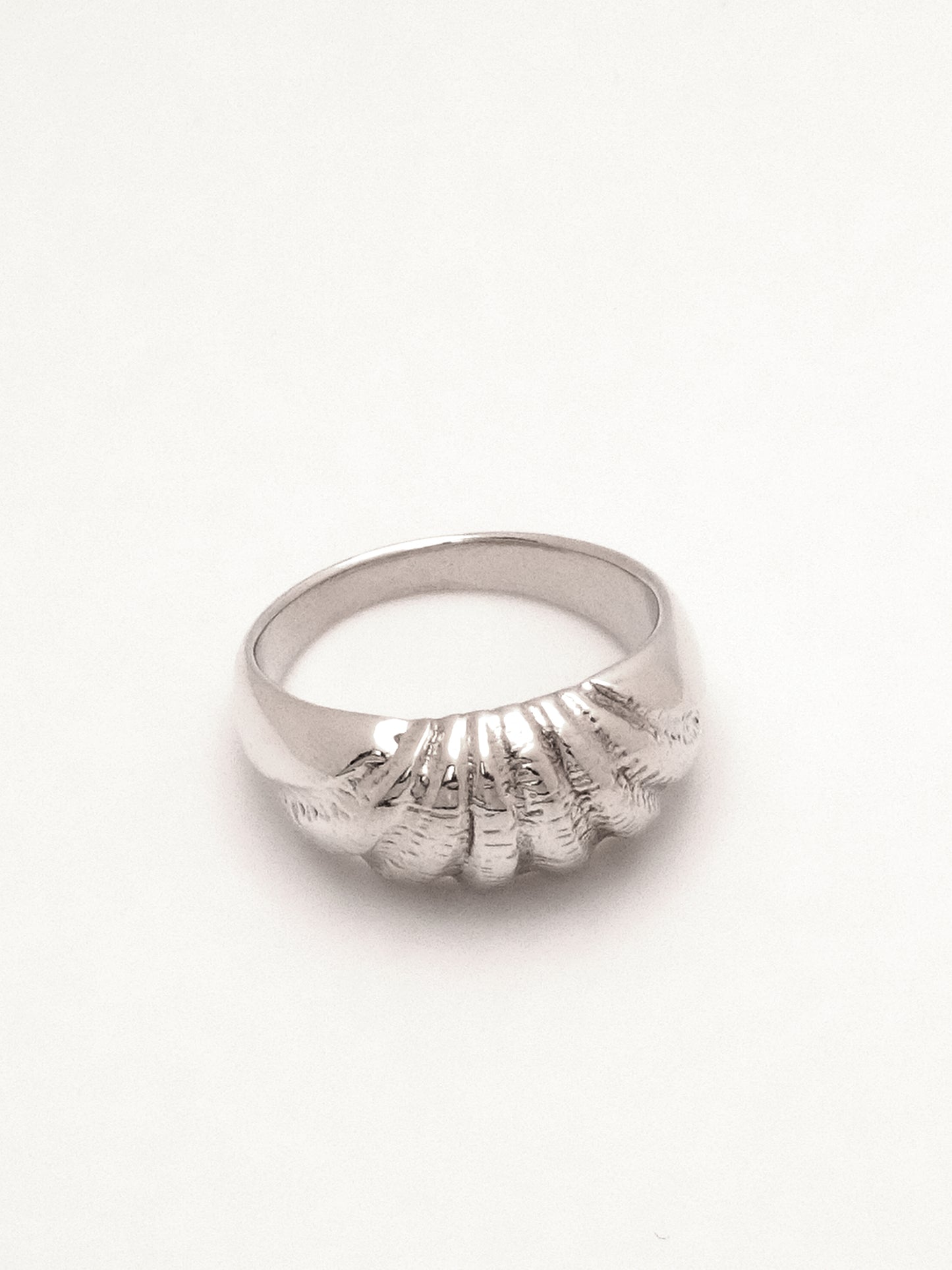 A sterling silver ring with a domed profile and intricate carving across the top mimicking the texture and radiating ridges of a seashell.  Shot lying flat from a three quarter topdown view to capture the ring's tapering form and the ridged texture. 