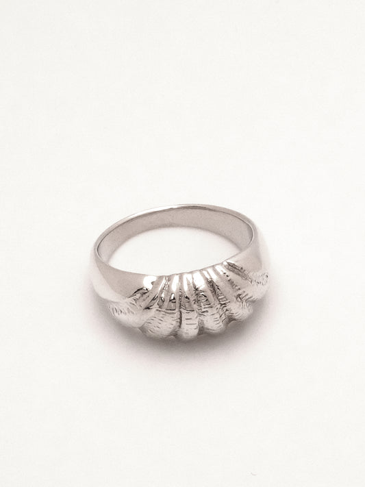 A sterling silver ring with a domed profile and intricate carving across the top mimicking the texture and radiating ridges of a seashell.  Shot lying flat from a three quarter topdown view to capture the ring's tapering form and the ridged texture. 